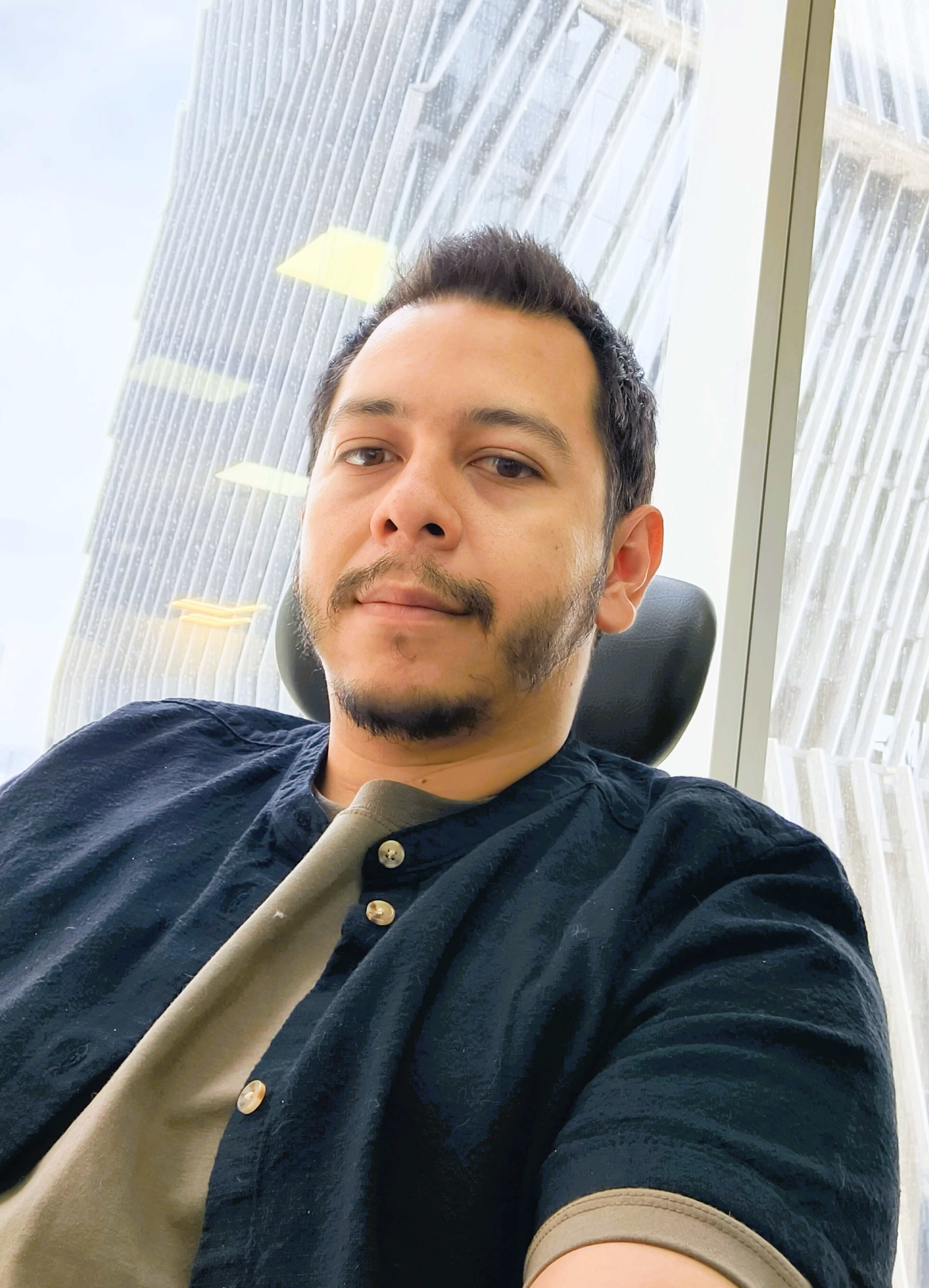 Joel Buenrostro en una fotografía casual sentado junto a una ventana, con camisa oscura y fondo arquitectónico moderno